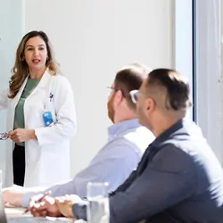 A female clinician points to a graph on the board as she explains the data represented to the seated male members.
