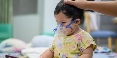 A toddler with an oxygen mask sits on a hospital bed and watches a phone.