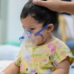 A toddler with an oxygen mask sits on a hospital bed and watches a phone.