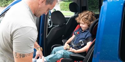 Boy with muscular dystrophy sitting in child safety seat in rear of family car, mature father fastening shoes in preparation of journey.