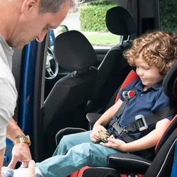 Boy with muscular dystrophy sitting in child safety seat in rear of family car, mature father fastening shoes in preparation of journey.