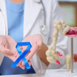 Close-up of female clinician holding a blue ribbon with both hands in front of her chest beside colon model on the table.