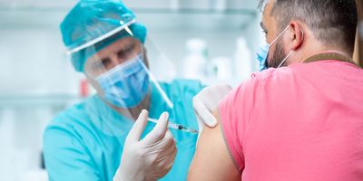 A clinician wearing a protective visor and surgical gloves injects a COVID-19 vaccine into a male patient's arm.