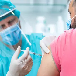 A clinician wearing a protective visor and surgical gloves injects a COVID-19 vaccine into a male patient's arm.