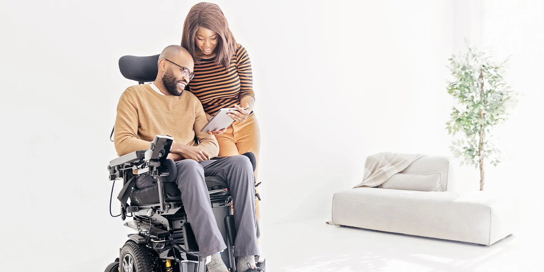 A bearded man in a wheelchair looks at a tablet device a woman standing beside him is holding.