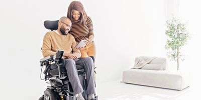 A bearded man in a wheelchair looks at a tablet device a woman standing beside him is holding.