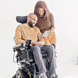 A bearded man in a wheelchair looks at a tablet device a woman standing beside him is holding.