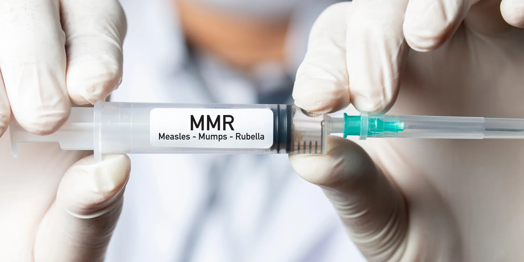 A pair of gloved hands holding a syringe labeled Measles, mumps, and rubella (MMR).