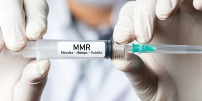 A pair of gloved hands holding a syringe labeled Measles, mumps, and rubella (MMR).