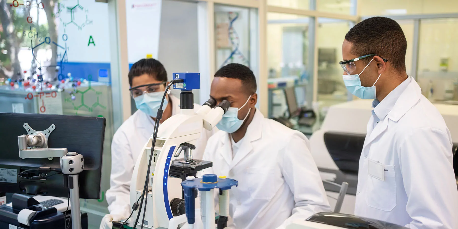 Photo of three clinical laboratory scientists wearing PPE examining clinical pathology samples under a microscope.