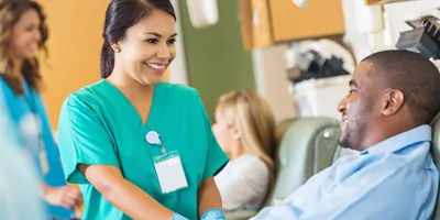 A smiling female nurse comforting a male patient lying on a hospital bed.