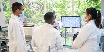  Photo of three clinical laboratory professionals wearing PPE standing in front of a glass window with green leaves in the background.