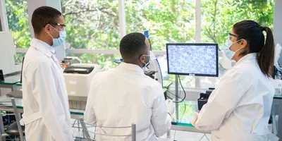 Photo of three clinical laboratory professionals wearing PPE standing in front of a glass window with green leaves in the background.