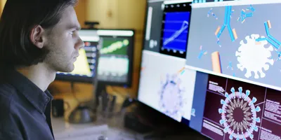 Stock photo of a young man working a large monitor displaying a variety of data & illustrations of viruses and antibodies