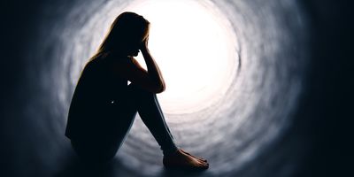 A silhouette of a woman sitting alone and holding her head in a dark tunnel with light at the other end.