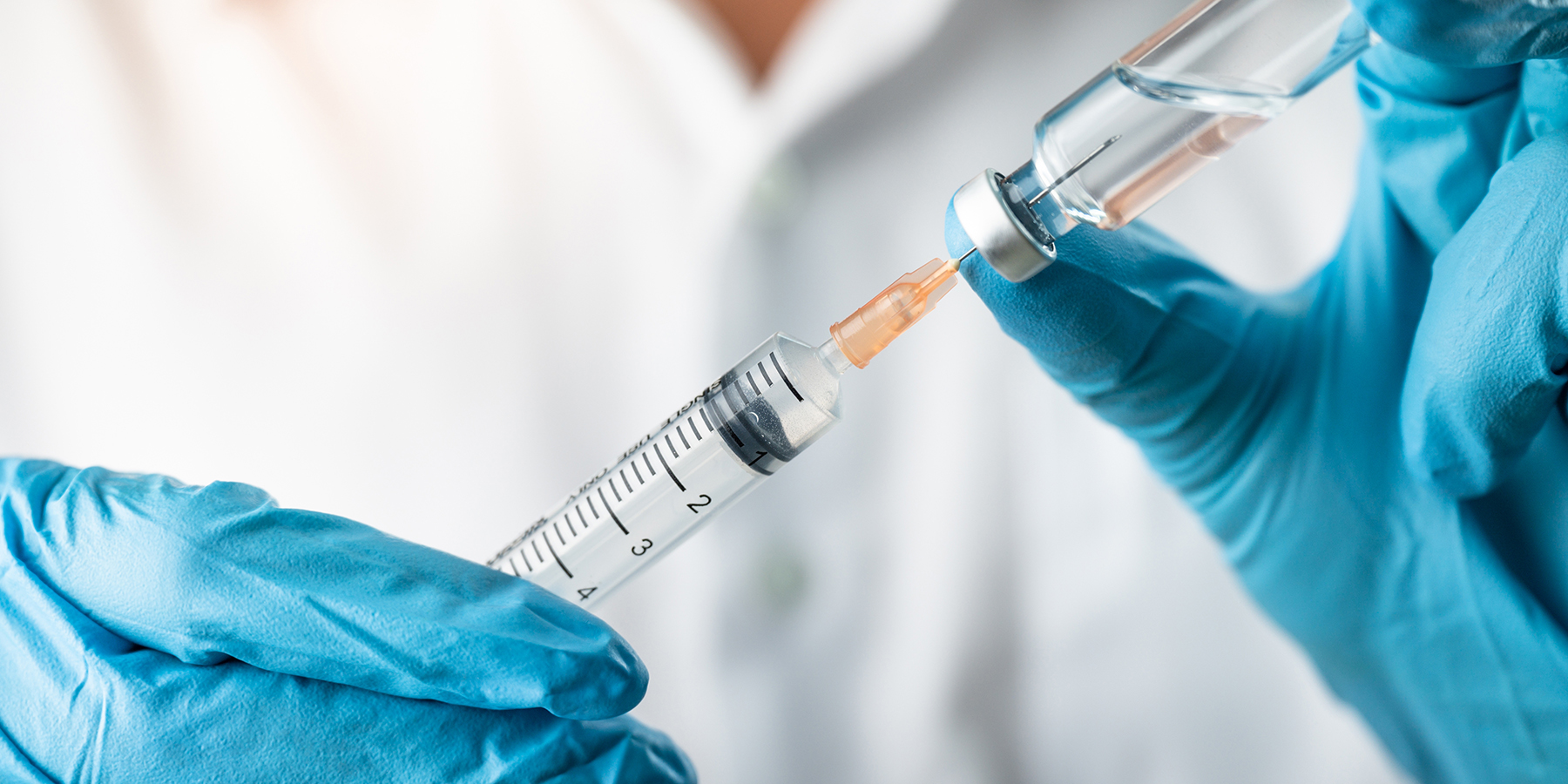 A pair of gloved hands load a clear, colorless liquid from a vial into a syringe.