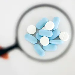 A hand holds a magnifying glass over a few capsules and tablets. 