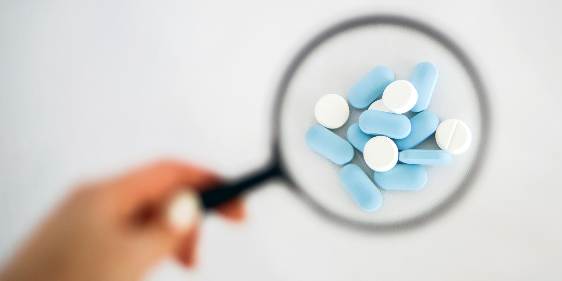 A hand holds a magnifying glass over a few capsules and tablets. 