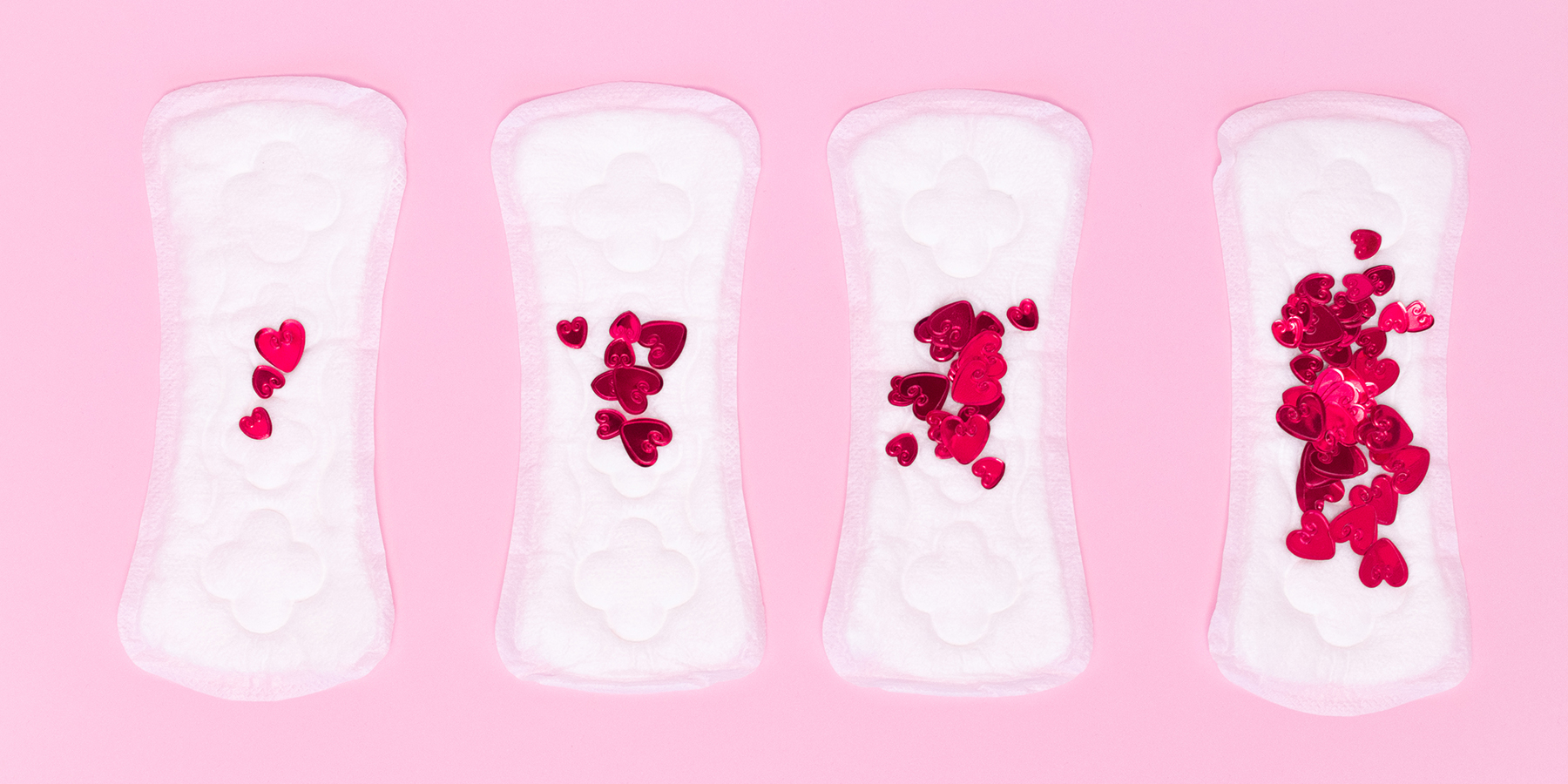Four sanitary napkins with an increasing number of heart-shaped confetti are arranged in tandem on a pink background.