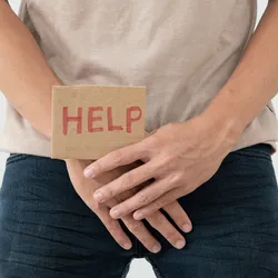 A man holds hands in front of his groin, showing a card with HELP written on it.