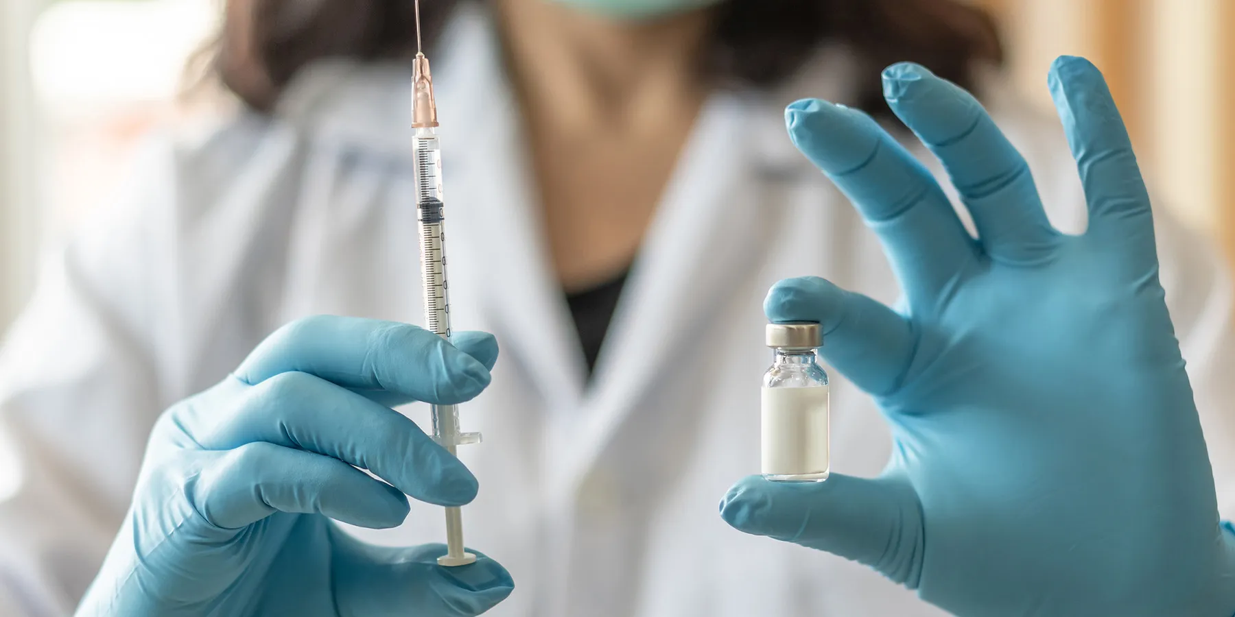 A clinician holds a syringe and a vaccine vial, each in a gloved hand.