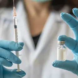 A clinician holds a syringe and a vaccine vial, each in a gloved hand.