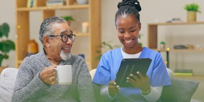 A female nurse-personal care worker smilingly shows a tab to a spectacled elderly man sitting holding a mug in his right hand.