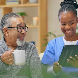 A female nurse-personal care worker smilingly shows a tab to a spectacled elderly man sitting holding a mug in his right hand.