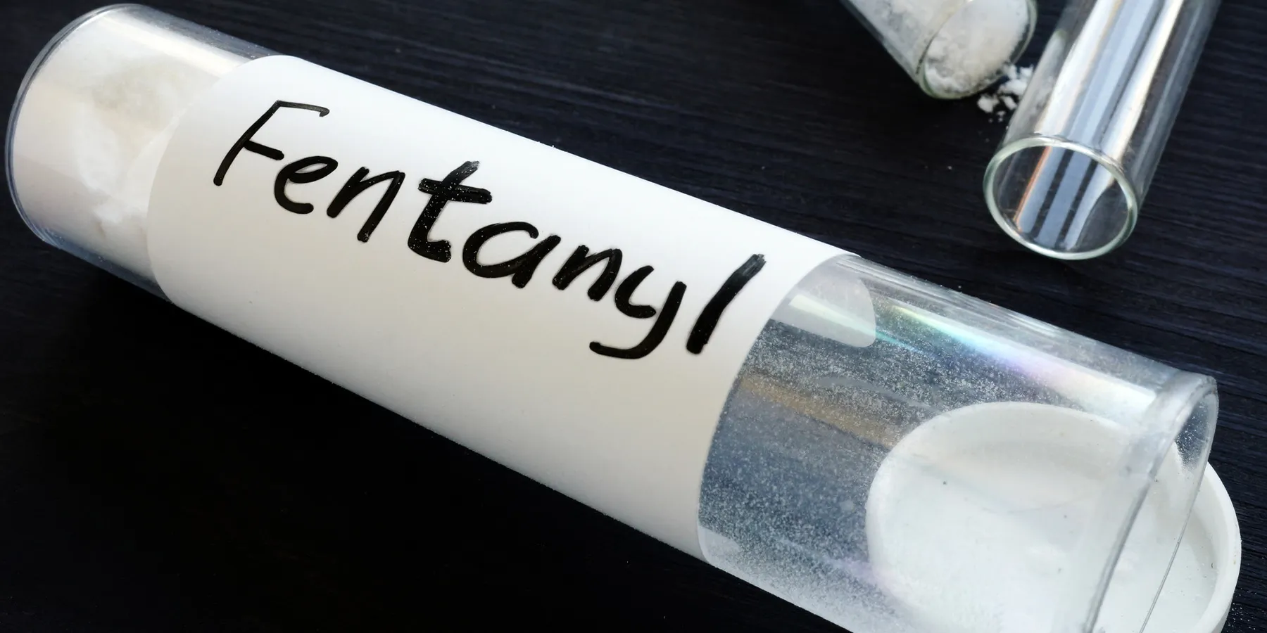 A transparent bottle with white powder, labeled Fentanyl, lays open with two more such bottles in the background.