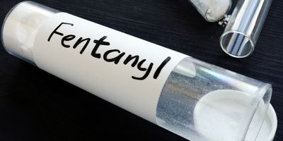 A transparent bottle with white powder, labeled Fentanyl, lays open with two more such bottles in the background.