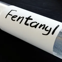 A transparent bottle with white powder, labeled Fentanyl, lays open with two more such bottles in the background.