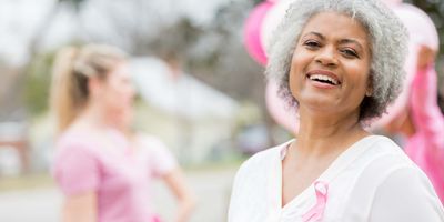 A cheerful senior African American woman, wearing a pink ribbon on the left side of her chest, smiles as she attends an outdoor breast cancer awareness event.