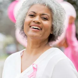 A cheerful senior African American woman, wearing a pink ribbon on the left side of her chest, smiles as she attends an outdoor breast cancer awareness event.