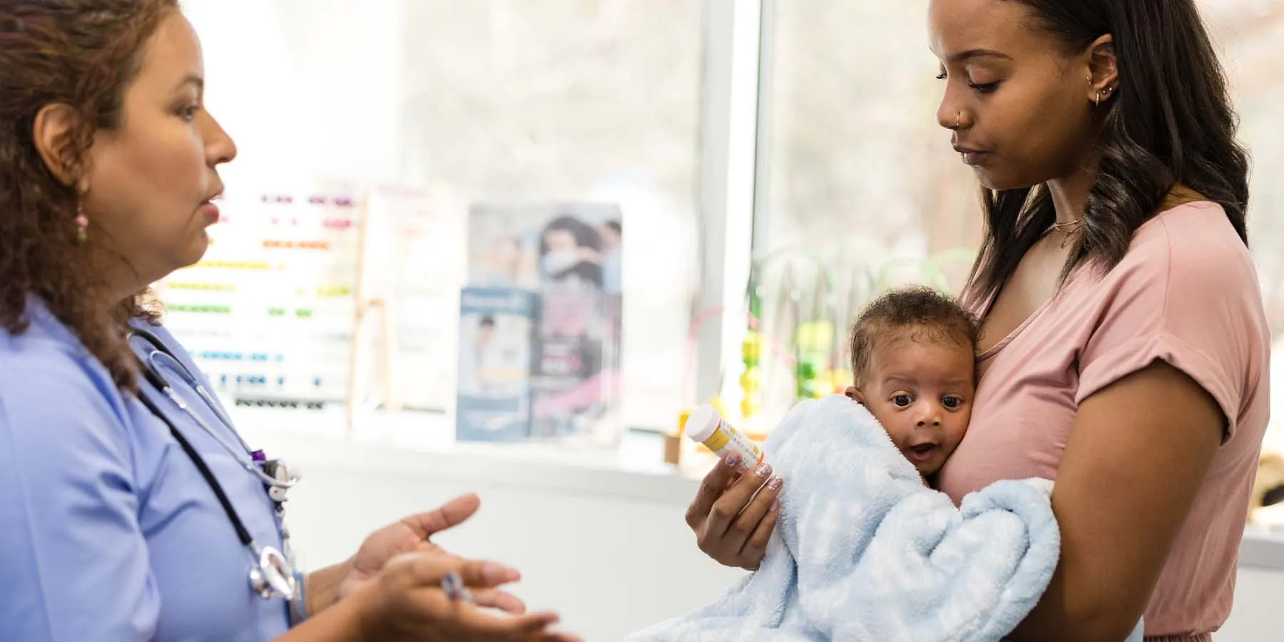 A female clinician gestures as she explains the dosage and side effects of a medication to a worried young mother holding a baby.