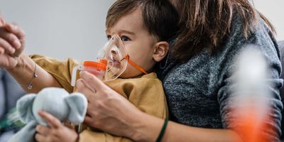 A baby boy takes inhalation treatment as he sits in his mother's lap, who is pointing at an object not in the frame.