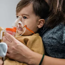A baby boy takes inhalation treatment as he sits in his mother's lap, who is pointing at an object not in the frame.