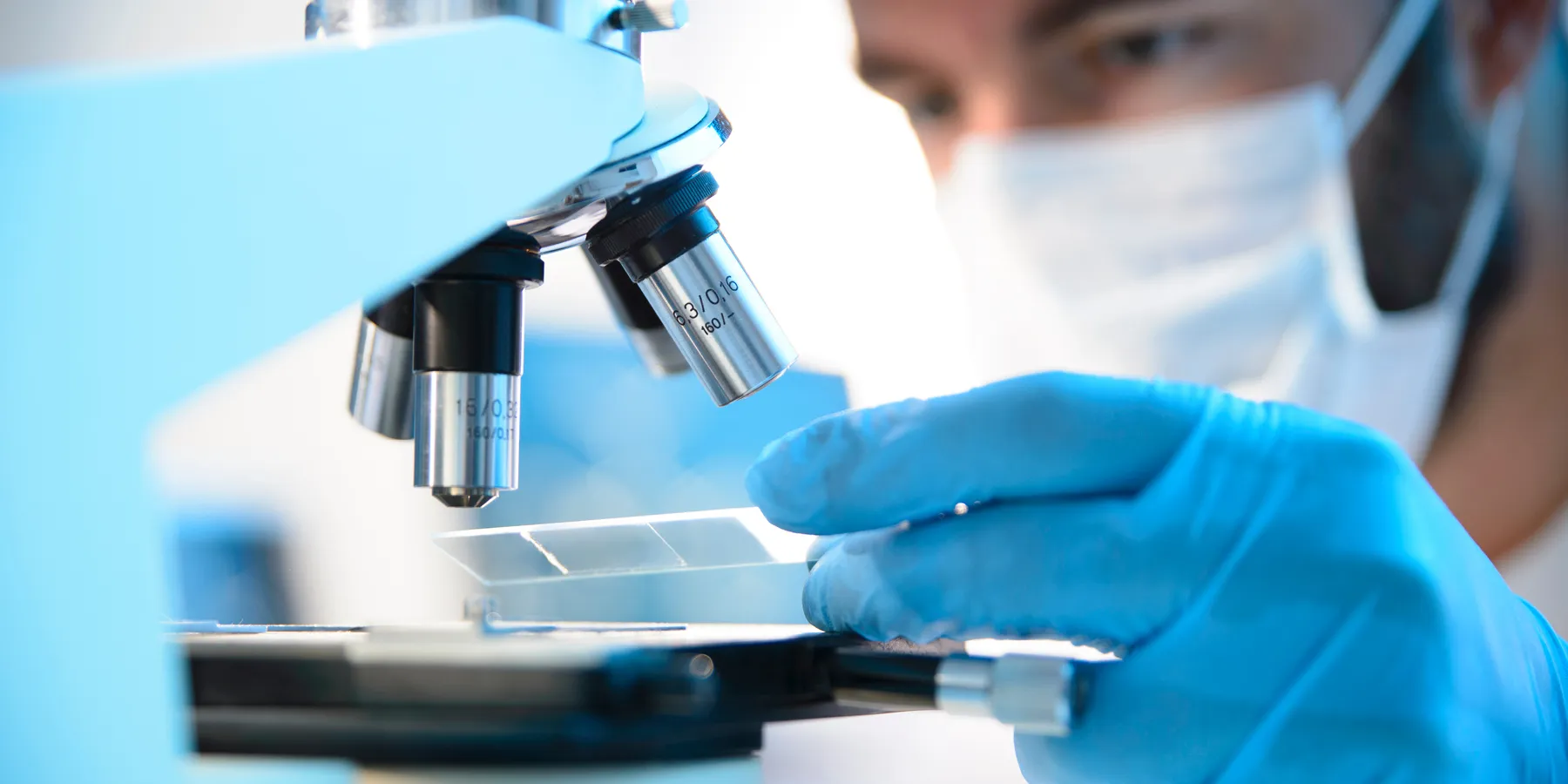 A male clinician wearing a mask inserts a prepared slide under a microscope.