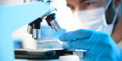 A male clinician wearing a mask inserts a prepared slide under a microscope.