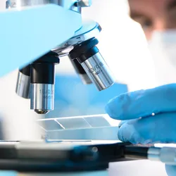 A male clinician wearing a mask inserts a prepared slide under a microscope.