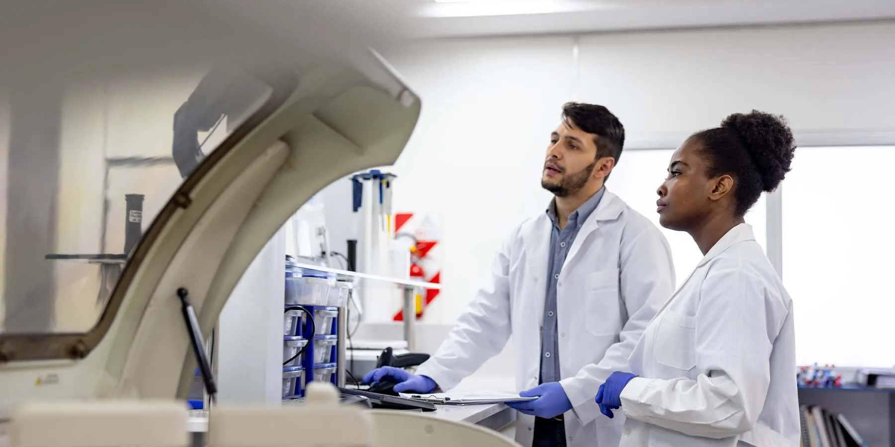 A male and a female scientist wearing PPE analyzing samples produced by automated equipment in a laboratory setting.