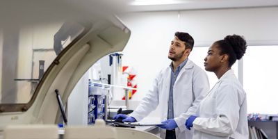 A male and a female scientist wearing PPE analyzing samples produced by automated equipment in a laboratory setting.