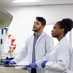 A male and a female scientist wearing PPE analyzing samples produced by automated equipment in a laboratory setting.