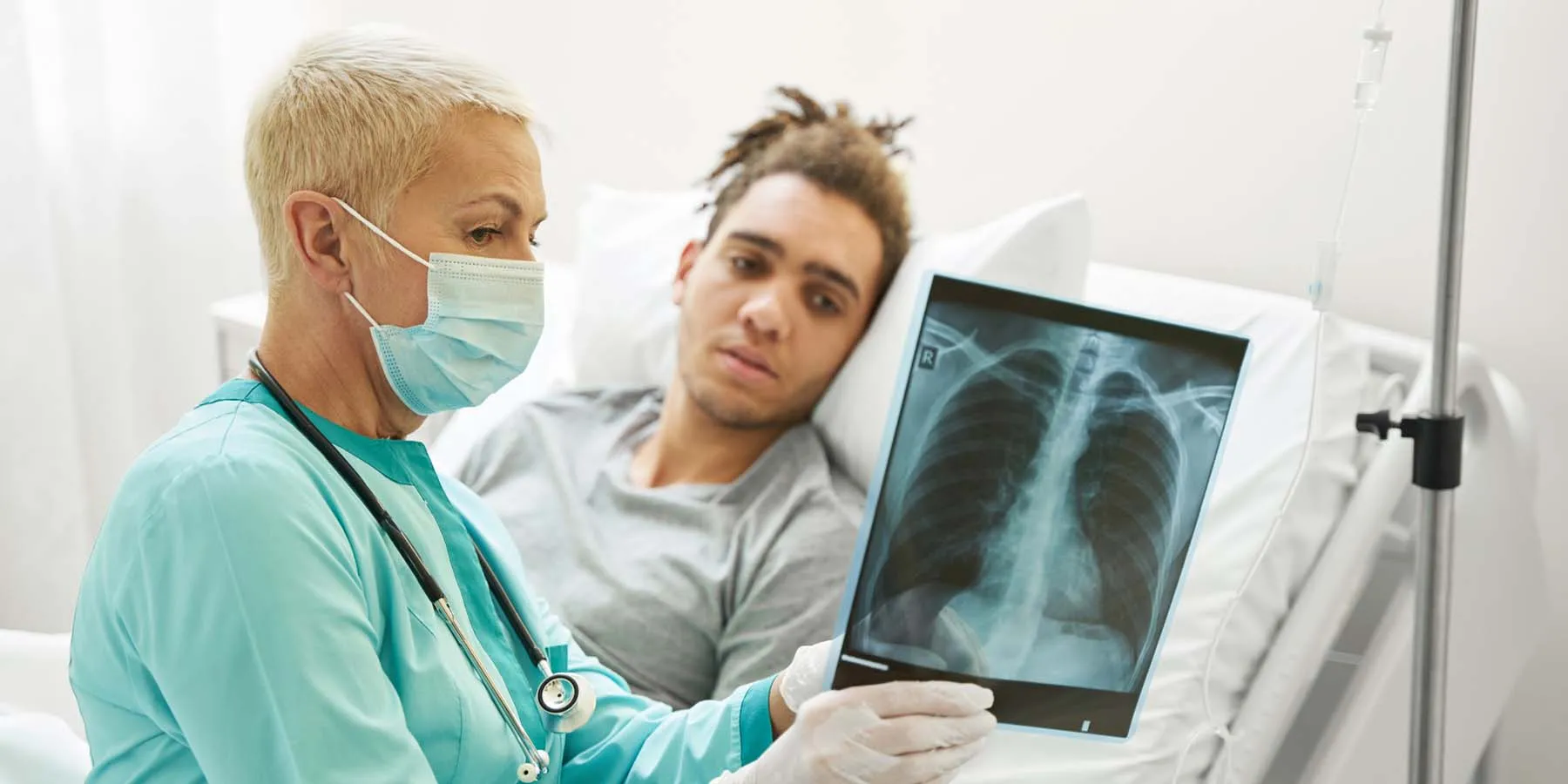 A female clinician explain a lung X-ray report to a male patient lying on the bed in a hospital room.