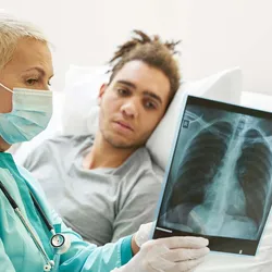 A female clinician explain a lung X-ray report to a male patient lying on the bed in a hospital room.