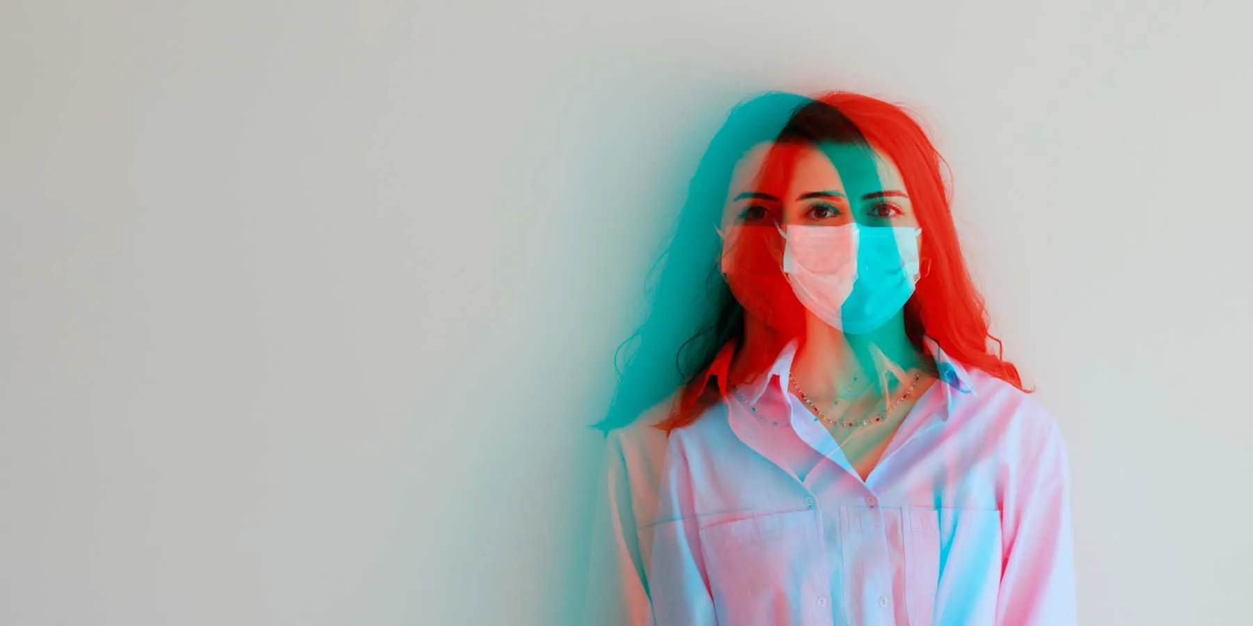 A young woman stands against a wall wearing a medical mask