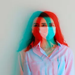 A young woman stands against a wall wearing a medical mask