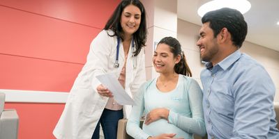 A female clinician shows a smiling pregnant couple a sheet of paper in an office.