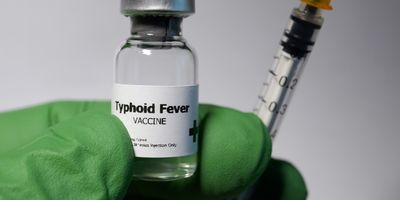 A gloved hand holds a syringe and a vial labeled Typhoid Fever Vaccine.