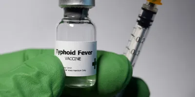 A gloved hand holds a syringe and a vial labeled Typhoid Fever Vaccine.