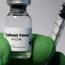 A gloved hand holds a syringe and a vial labeled Typhoid Fever Vaccine.